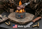 A small cylindrical camping stove with visible flames holds a piece of meat over a stone surface, with a person in camouflage gloves holding tongs above it. Text on the image reads 'ALL NATURAL AND / SAFE FOR COOKING' and 'Made in USA', with a BBUSA logo on the stove.